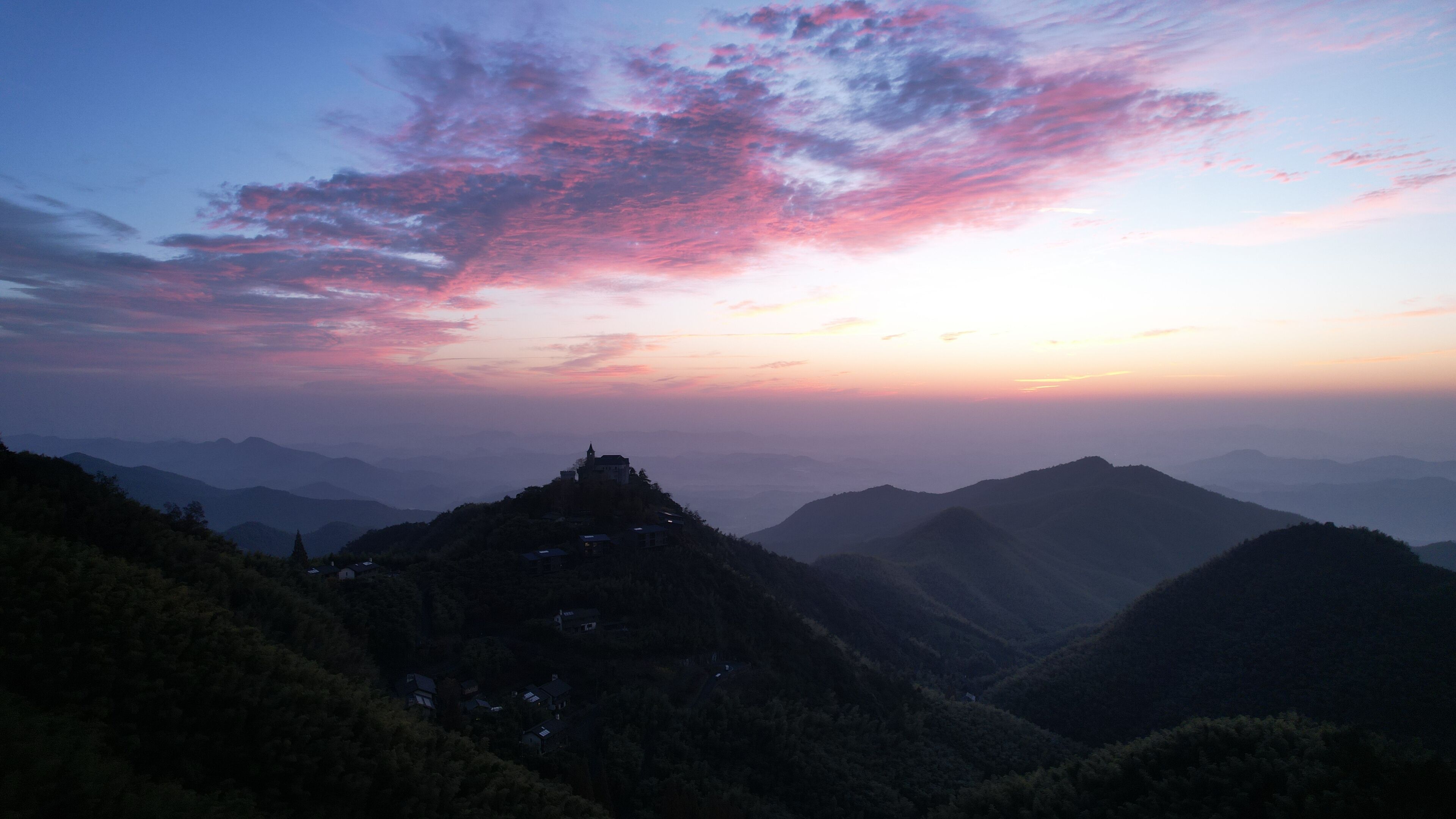 A mountainous landscape with a sunset, featuring a prominent peak and scattered buildings.