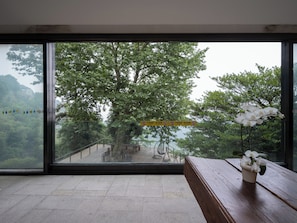 A room with a large window overlooking a forested area and a wooden bench with a potted orchid.