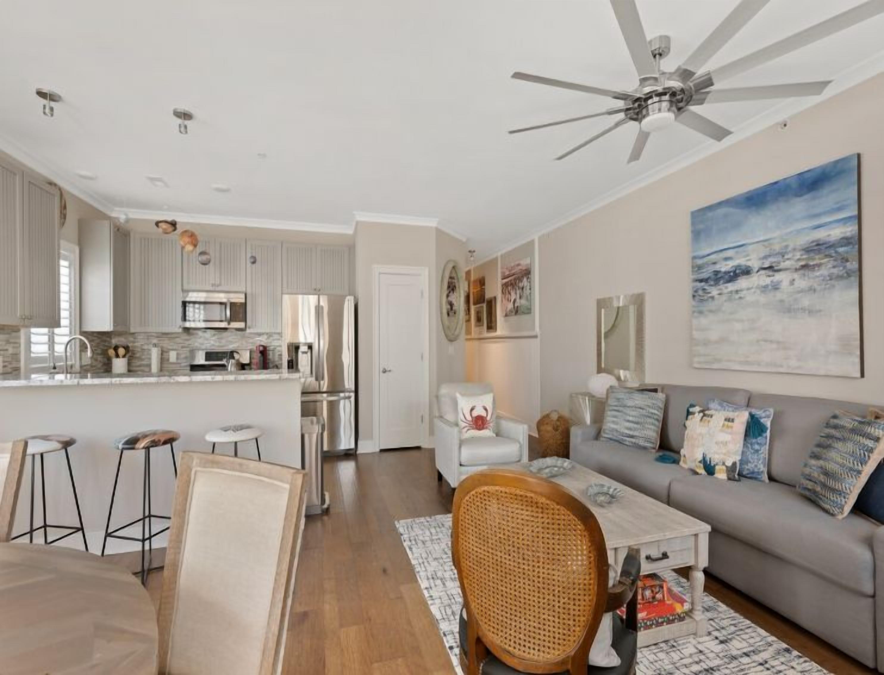 Une cuisine moderne dotée d’un îlot central, un coin repas avec des tabourets, un salon avec un canapé et des coussins, et un ventilateur de plafond.