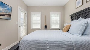Un dormitorio con cama, dos ventanas con persianas, una puerta y un cuadro colgado en la pared.