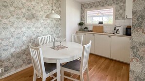 Un comedor con una mesa redonda, cuatro sillas, una lámpara colgante y una cocina con ventana y electrodomésticos en la encimera.