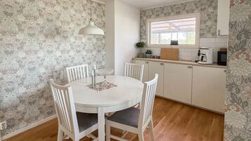 A dining area with a round table, four chairs, a hanging light fixture, and a kitchen with a window and countertop appliances.