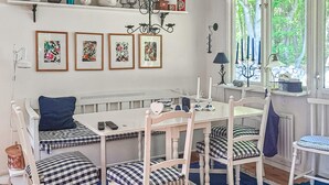Un comedor con una mesa blanca, cuatro sillas con cojines a cuadros, un banco con cojín azul y un ventanal con vistas a árboles.
