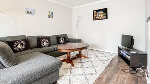 En stue med en grå sofa, et rundt træbord, et hvidt tæppe og et tv-møbel med et fladskærms-tv.