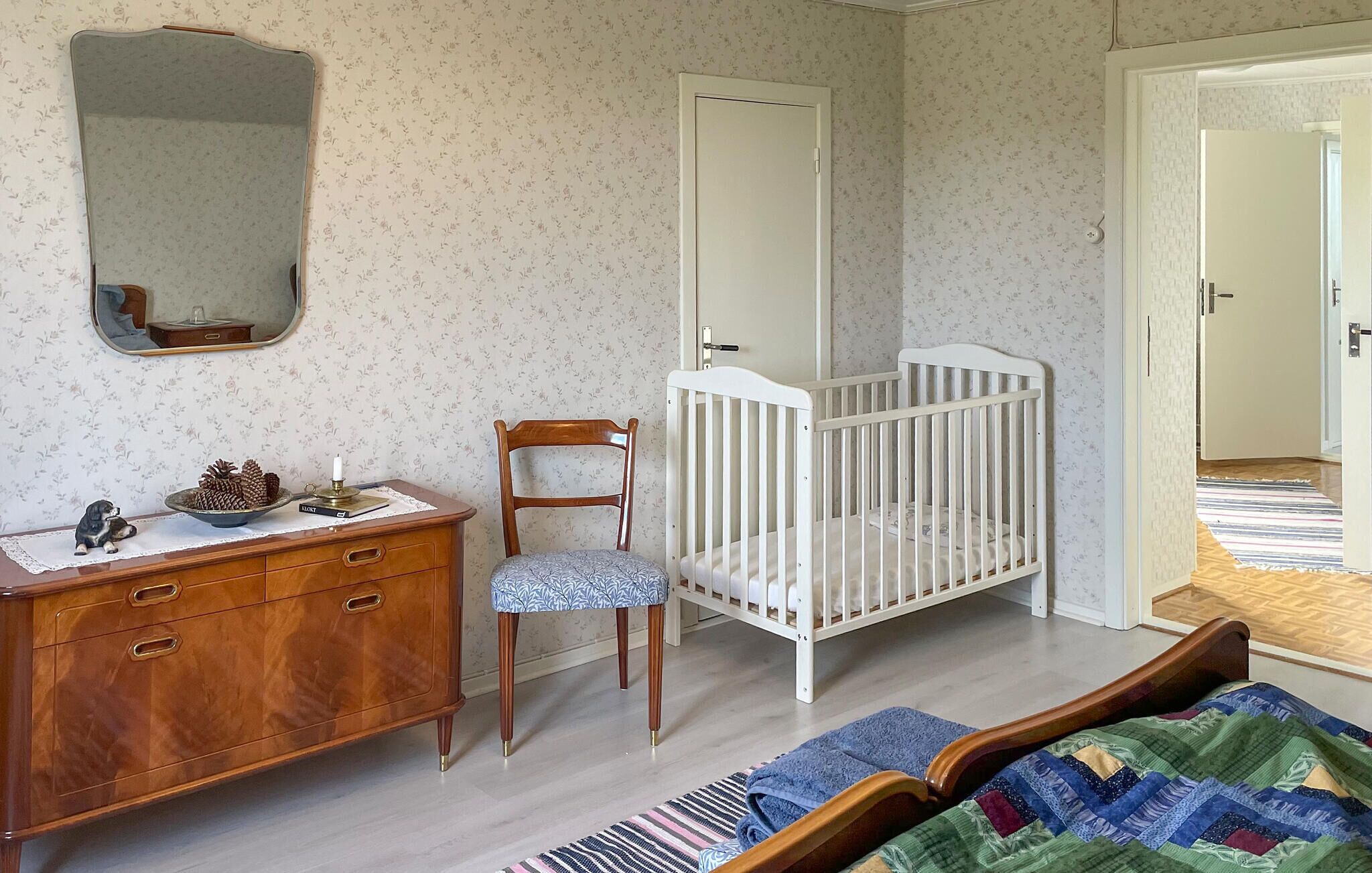 Een kamer met een houten dressoir, een wit wiegje, een stoel en een bed met een sprei.