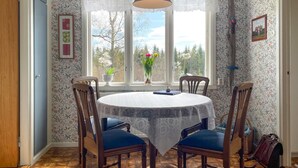 Ein Essbereich mit einem runden Tisch, vier Stühlen, einer Spitzenleinentischdecke, einer Hängeleuchte und einem Fenster mit Blick auf Bäume und ein Haus.