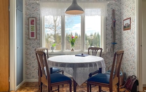 Ein Essbereich mit einem runden Tisch, vier Stühlen, einer Spitzenleinentischdecke, einer Hängeleuchte und einem Fenster mit Blick auf Bäume und ein Haus.