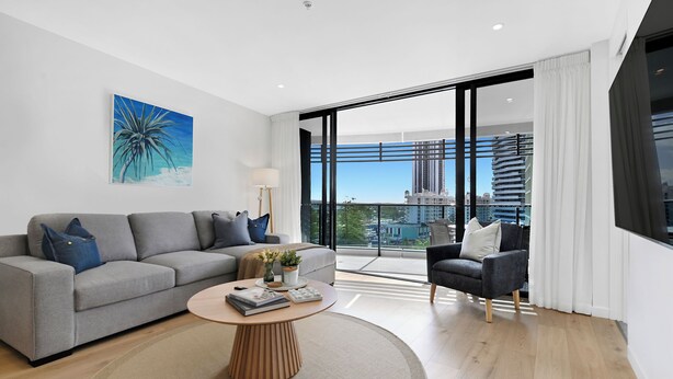 A modern living room with a grey sofa, a round coffee table, a black armchair, and a large window with a city view. - Beach Holiday at Oracle Level 7 (Gold Coast)