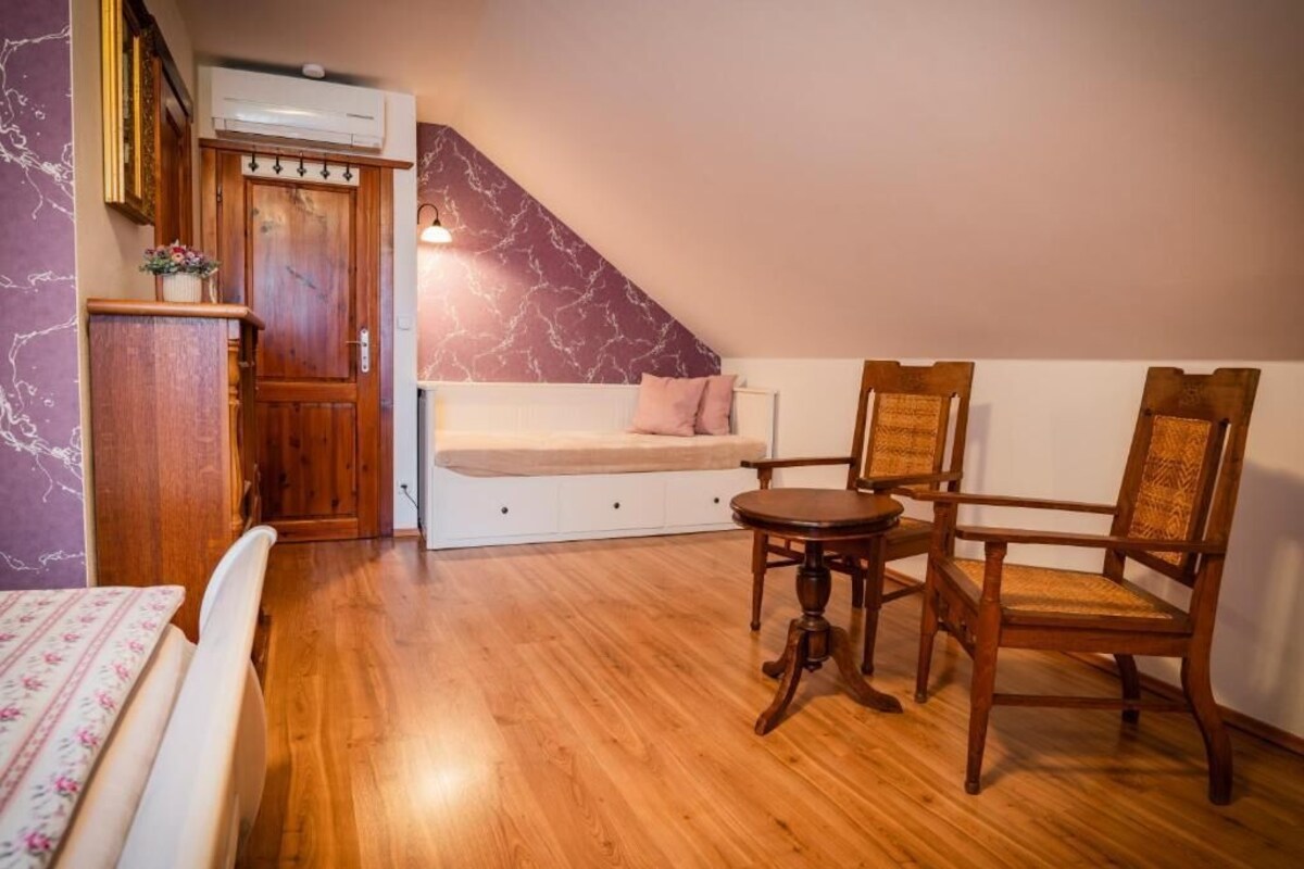 A room with a wooden floor, a bed with a floral bedspread, a bench with a cushion, a small round table, and two wooden chairs.
