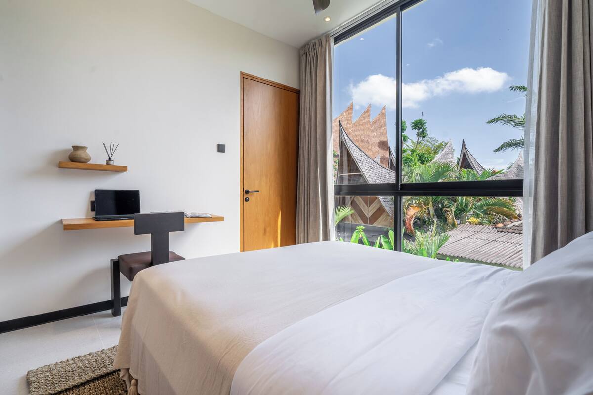 Una habitación de hotel moderna con una cama grande, un escritorio con una computadora portátil, un estante pequeño con objetos decorativos, y una ventana que ofrece vistas a la arquitectura tradicional y al verdor.
