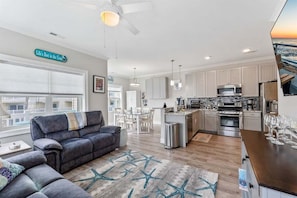A modern living room with a gray sofa, a star-patterned rug, and a kitchen with white cabinets and stainless steel appliances. - Residence 201S at The Sandcastle Condominiums (Wildwood Crest)