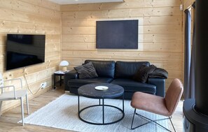 Ein gemütliches Blockhaus-Interieur mit einem schwarzen Ledersofa, einem runden Couchtisch, einem einzelnen Sessel, einem Flachbildfernseher und einem Kamin.