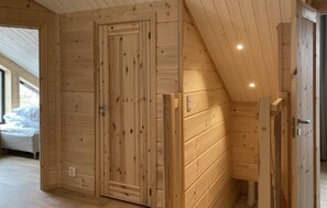 Ein Holzinnenraum mit schräg verlaufender Decke, einer Tür und einem Bett im angrenzenden Zimmer.