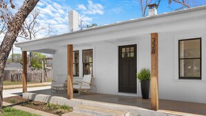 Ein weißes Einfamilienhaus mit schwarzem Eingangstür, zwei Fenstern und einer Veranda mit einem Stuhl.
