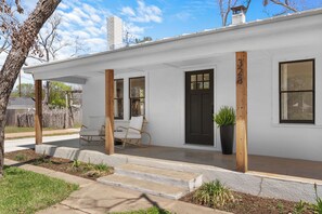 Ein weißes Einfamilienhaus mit schwarzem Eingangstür, zwei Fenstern und einer Veranda mit einem Stuhl.