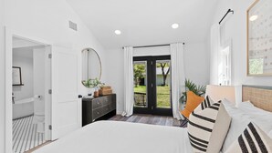 Ein Schlafzimmer mit Bett, Kommode, Spiegel, Fenster mit Vorhängen und Blick ins Freie.