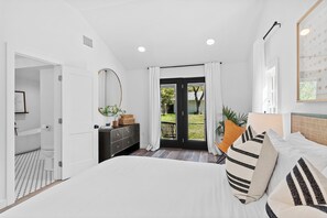 Ein Schlafzimmer mit Bett, Kommode, Spiegel, Fenster mit Vorhängen und Blick ins Freie.