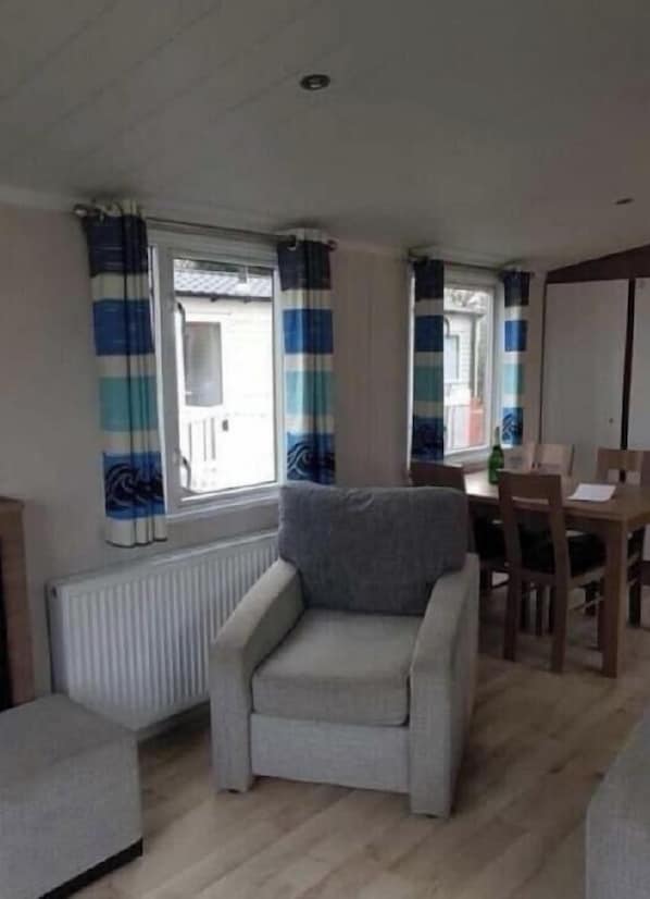 Ein Zimmer mit grauem Sessel, einem Esstisch mit Stühlen, einem Heizkörper und Fenstern mit gestreiften Vorhängen.