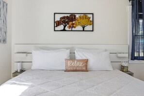 Ein Bett mit weißer Bettwäsche, ein Kissen mit dem Aufdruck 'Relax You're on beach time' und ein Wandbild von Herbstbäumen.