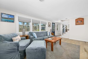 Ein Wohnzimmer mit einer Eckcouch, einem Holztisch und einem großen Fenster mit Blick auf Bäume.