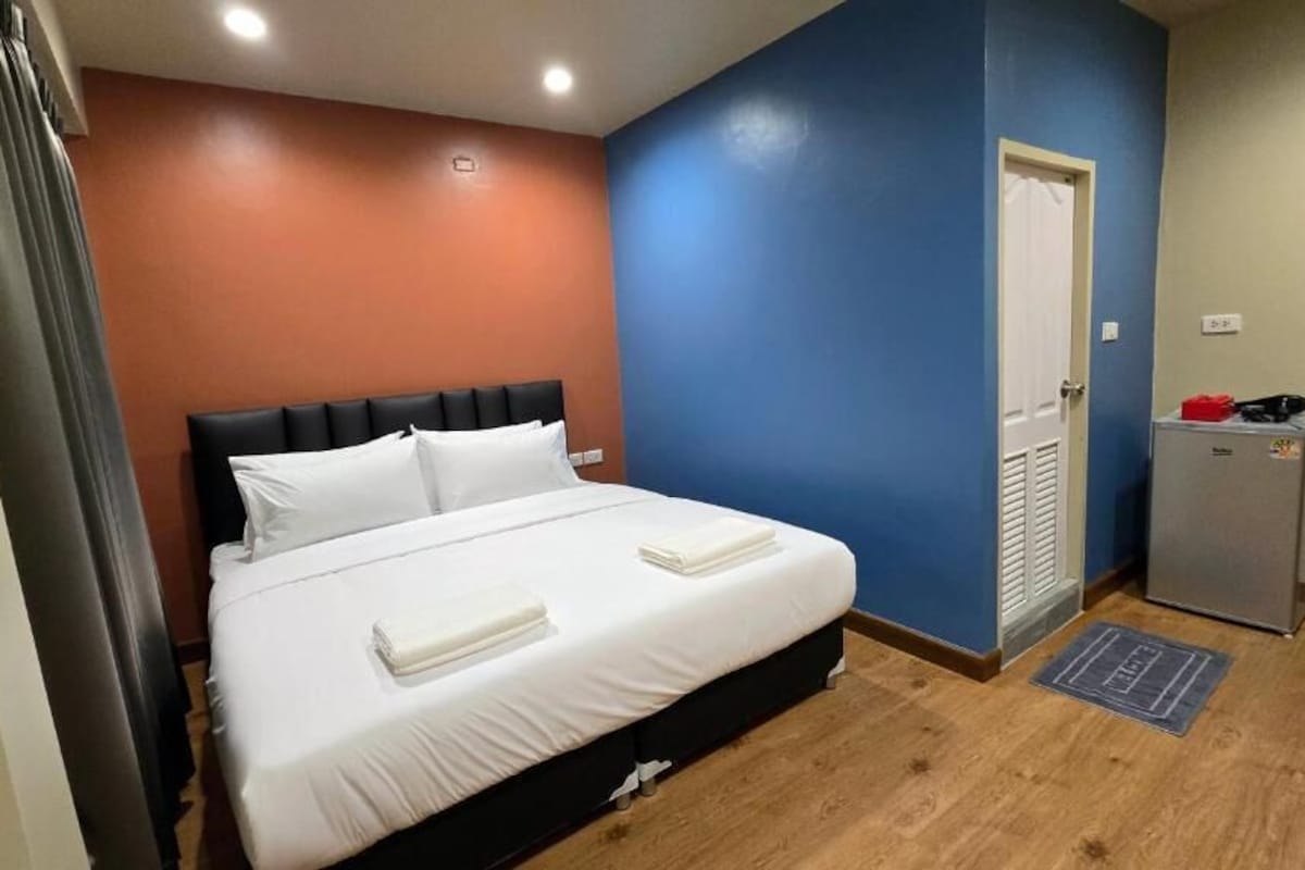 A bedroom with a bed, a door, a small refrigerator, and a wooden floor.