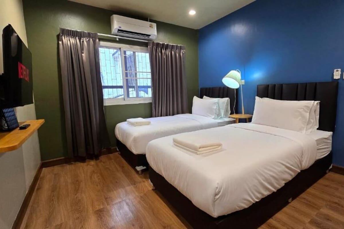 A hotel room with two beds, a TV, a window with curtains, a lamp, and an air conditioning unit.