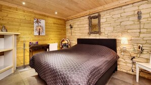 Une chambre avec un plafond en bois, un mur en pierre, un lit recouvert d’une courtepointe à motifs, un bureau avec une lampe et une étagère.