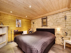 Ein Schlafzimmer mit Holzboden, Steinwand, einem Bett mit gemustertem Überwurf, einem Schreibtisch mit Lampe und einem Regal.