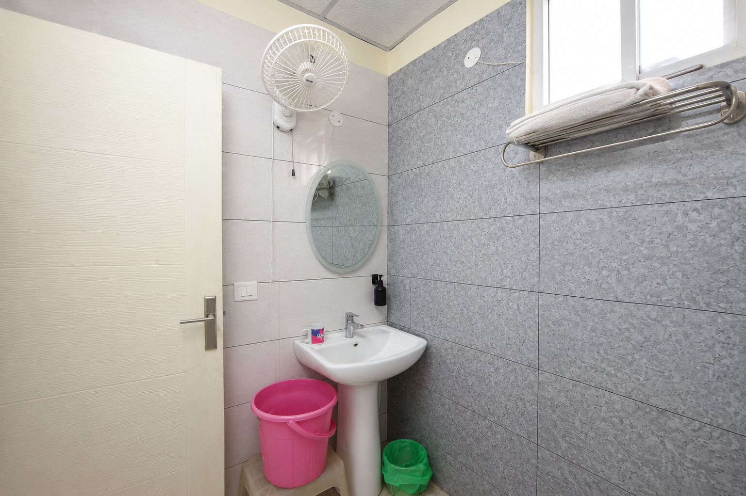 Ein Badezimmer mit einem weißen Waschbecken, einem runden Spiegel, einem weißen Ventilator, einem Handtuchhalter und einem Fenster.