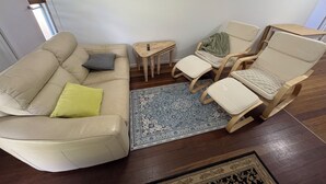 En stue med en beige sofa, to træstole, et lille træbord og en blå måtte med et mønster.