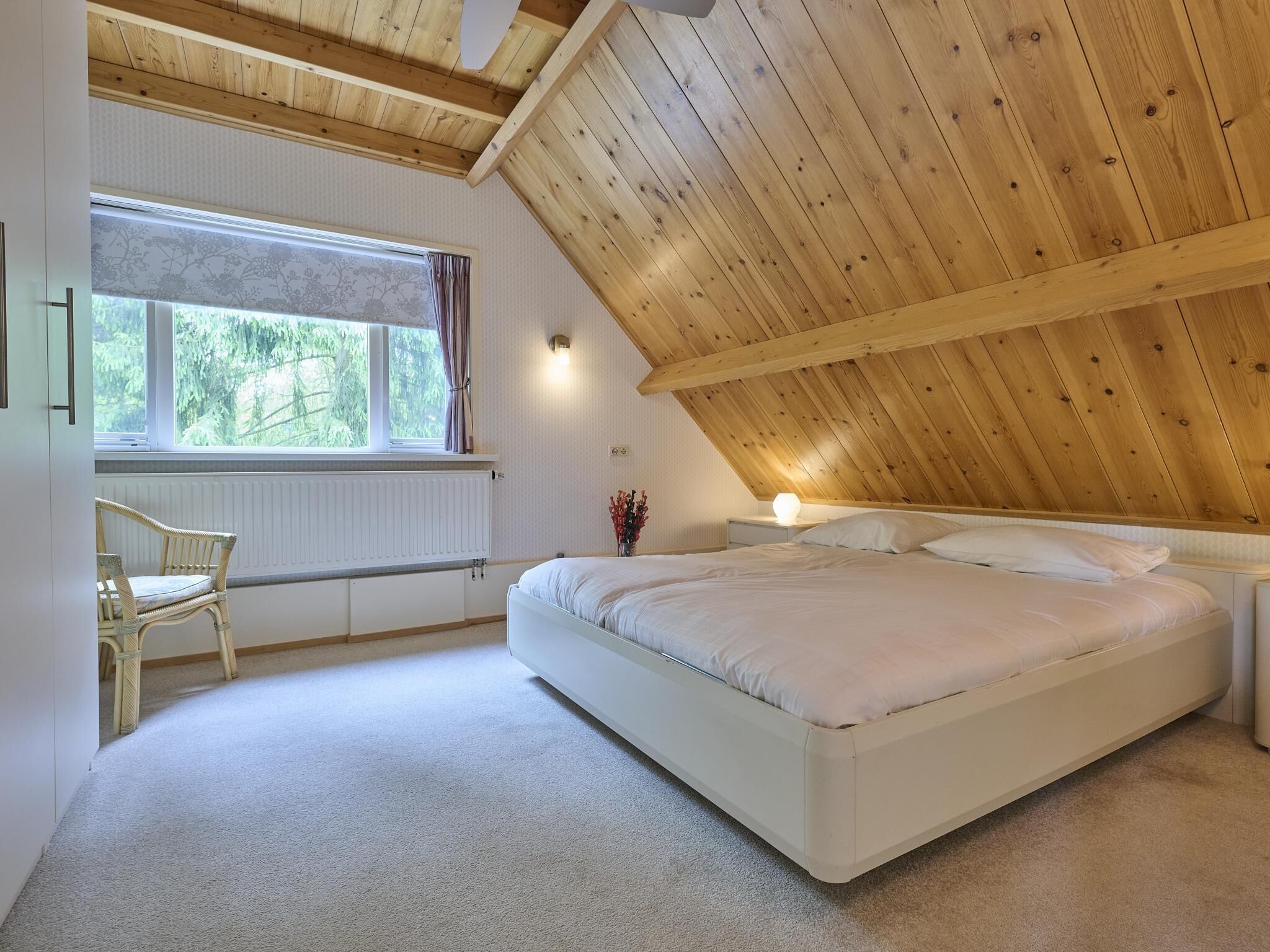 A bedroom with a sloped ceiling, a bed, a chair, a window with a view of greenery, and a radiator.