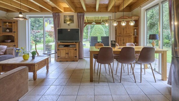 A spacious living area with a wooden ceiling, a dining table with chairs, a TV on a wooden stand, and large windows offering a view of greenery. - Home in a private park with a spacious garden (Epe)