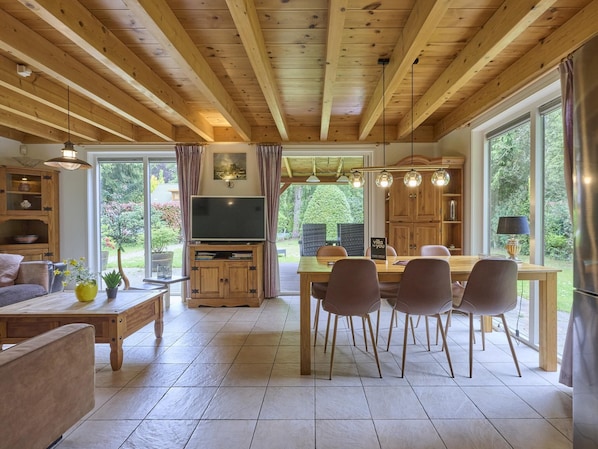 A spacious living area with a wooden ceiling, a dining table with chairs, a TV on a wooden stand, and large windows offering a view of greenery. - Home in a private park with a spacious garden (Epe)