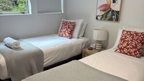 A hotel room with two single beds, a nightstand with a lamp, a painting on the wall, and a window with blinds. - Bedroom (Jack's Point)