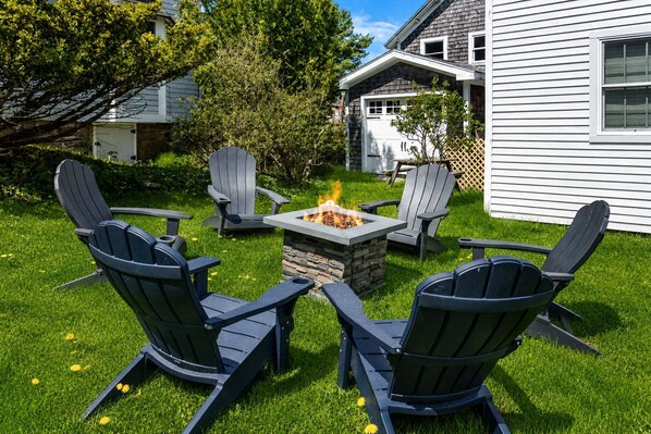 A backyard with a stone fire pit surrounded by four Adirondack chairs.