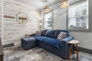 A room with a sectional sofa, a small stool, a wall-mounted mirror, and two windows with blinds.
