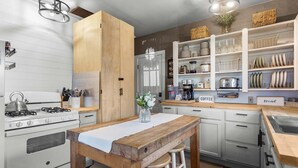 A kitchen with white cabinets, a wooden table, stools, a stove, and a coffee station. - Winter Harbor Haven - NEW! (Winter Harbor)