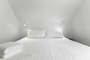 A white, high-ceilinged room with a bed, two pillows, and a folded towel.