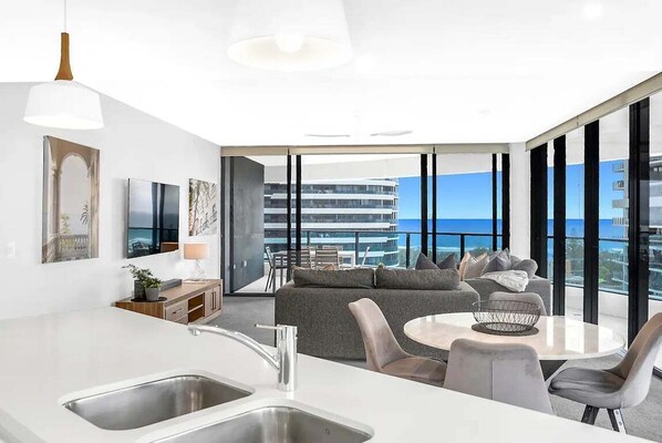 A modern kitchen with a large island, a dining area with a round table, and a sofa with a view of the ocean. - Relax at the Beach at Oracle Level 10 (Gold Coast)