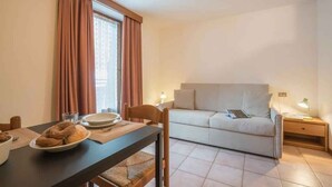 Ein Zimmer mit Esstisch, einer Couch, einem Fenster mit Vorhängen und einem Nachttisch mit Lampe.