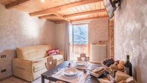 Ein gem ütliches Zimmer mit Holzbalkendecke, einer Couch, einem Esstisch mit Tellern, Gläsern, Brot und einer Weinflasche.