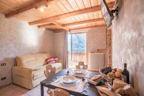 Ein gemütliches Zimmer mit Holzbalkendecke, einer Couch, einem Esstisch mit Tellern, Gläsern, Brot und einer Weinflasche.