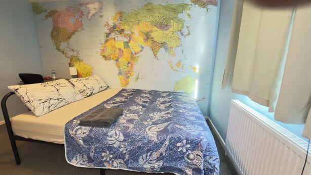 A bedroom with a bed, pillows, a map on the wall, a radiator, and a window. - Room-budget Friendly Private Room - Your (Wellingborough)