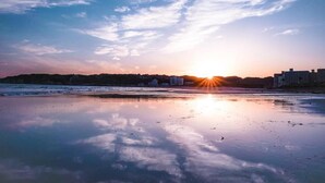 A beach with the sun setting over the horizon, calm water, and buildings in the background. - DEAR MOM (Onjuku)