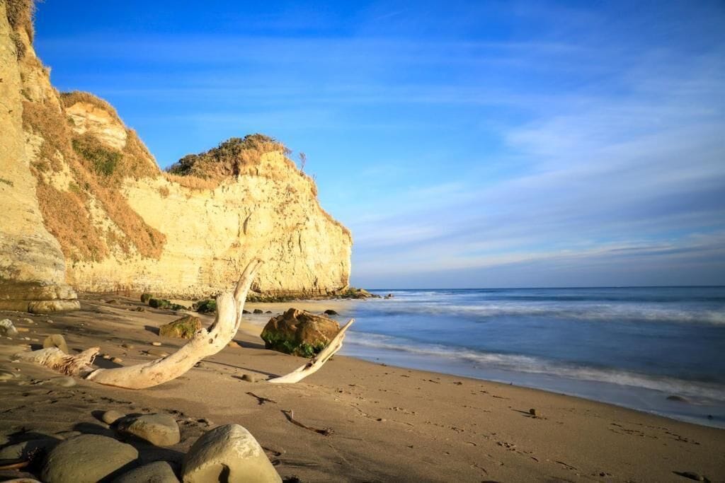 Une plage avec une grande formation rocheuse, un morceau de bois flotté et un ciel dégagé.