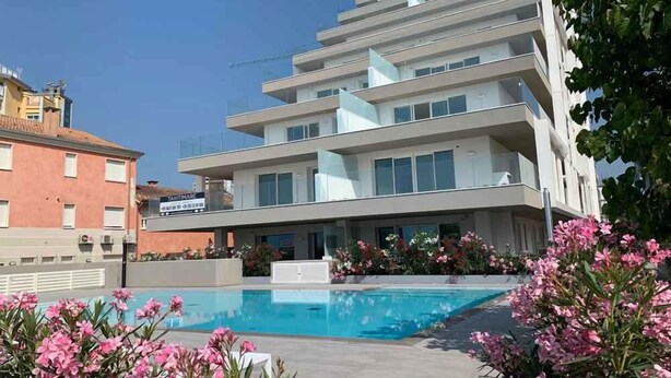 A modern multi-story building with a swimming pool and pink flowers in the foreground. - Holiday flat with balcony (Lido di Jesolo)