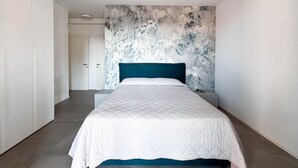 A bedroom with a bed, a nightstand, a wall with a floral pattern, and a door leading to another room. - Holiday flat with balcony (Lido di Jesolo)