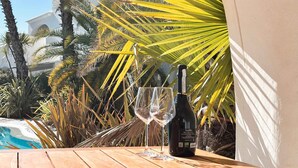 A bottle of wine and two glasses on a wooden table with a pool and palm trees in the background. - Holiday apartment with communal pool (Lido di Jesolo)
