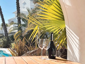A bottle of wine and two glasses on a wooden table with a pool and palm trees in the background. - Holiday apartment with communal pool (Lido di Jesolo)