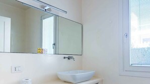 A bathroom with a white sink, a mirror, a window, and a toilet paper dispenser. - Holiday apartment with communal pool (Lido di Jesolo)